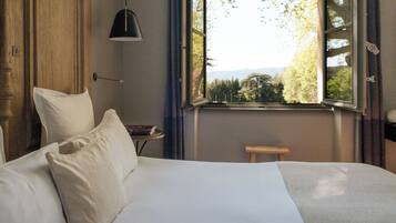 Une chambre à coucher avec un lit, une table de chevet sur laquelle se trouve un livre, une lampe et une fenêtre donnant sur des arbres et des montagnes.