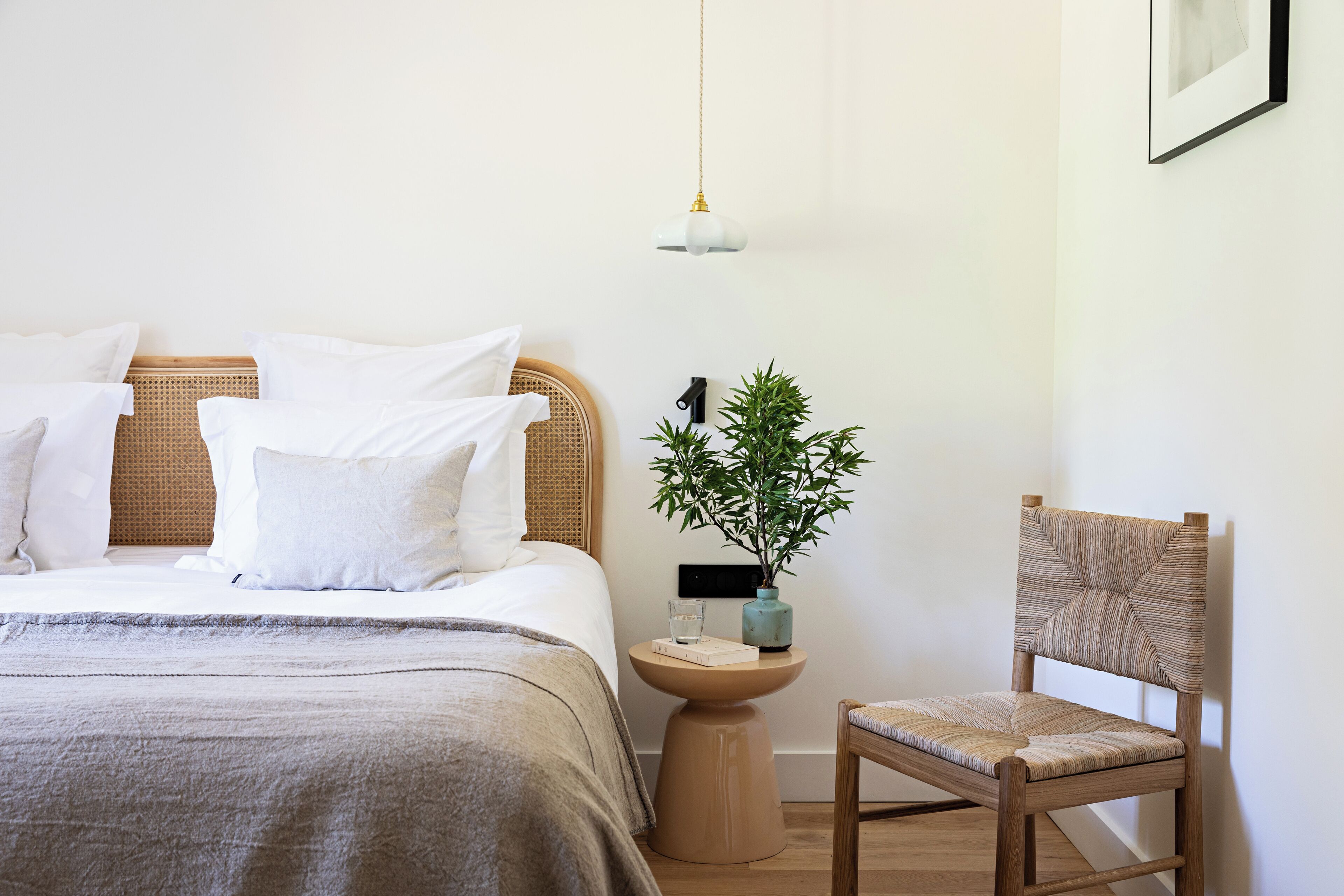 A neatly made bed with white linens, a bedside table with a glass and a book, a potted plant, and a wooden chair.