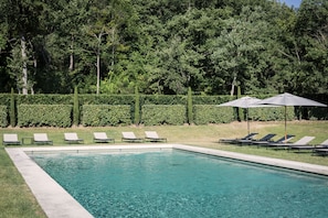 Una piscina rectangular con agua cristalina y azul, rodeada por un seto bajo y una fila de sillones de descanso.