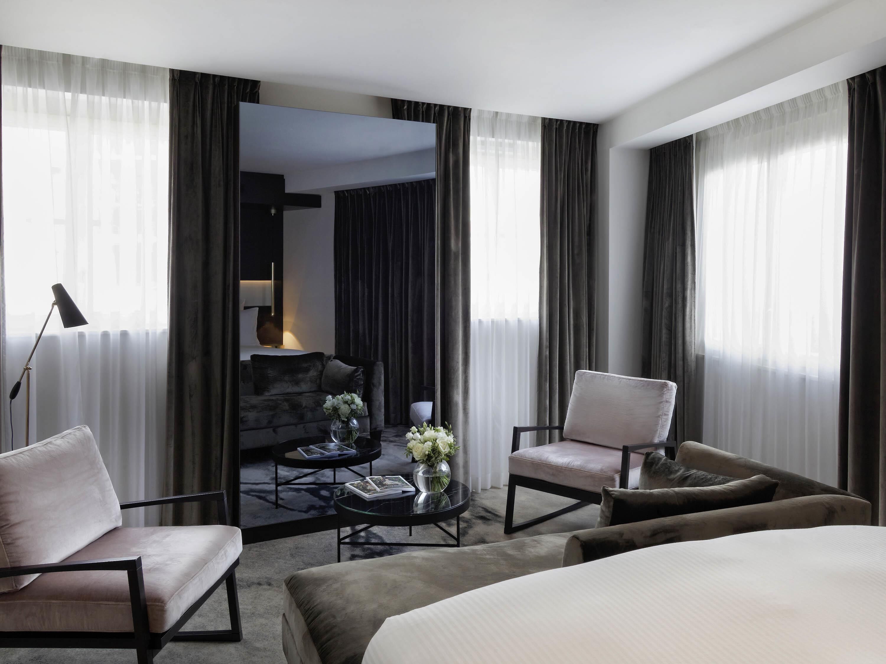 Une chambre d’hôtel avec un lit, deux fauteuils, une table basse et un canapé. Il y a un miroir au mur et une lampe sur la table de chevet.
