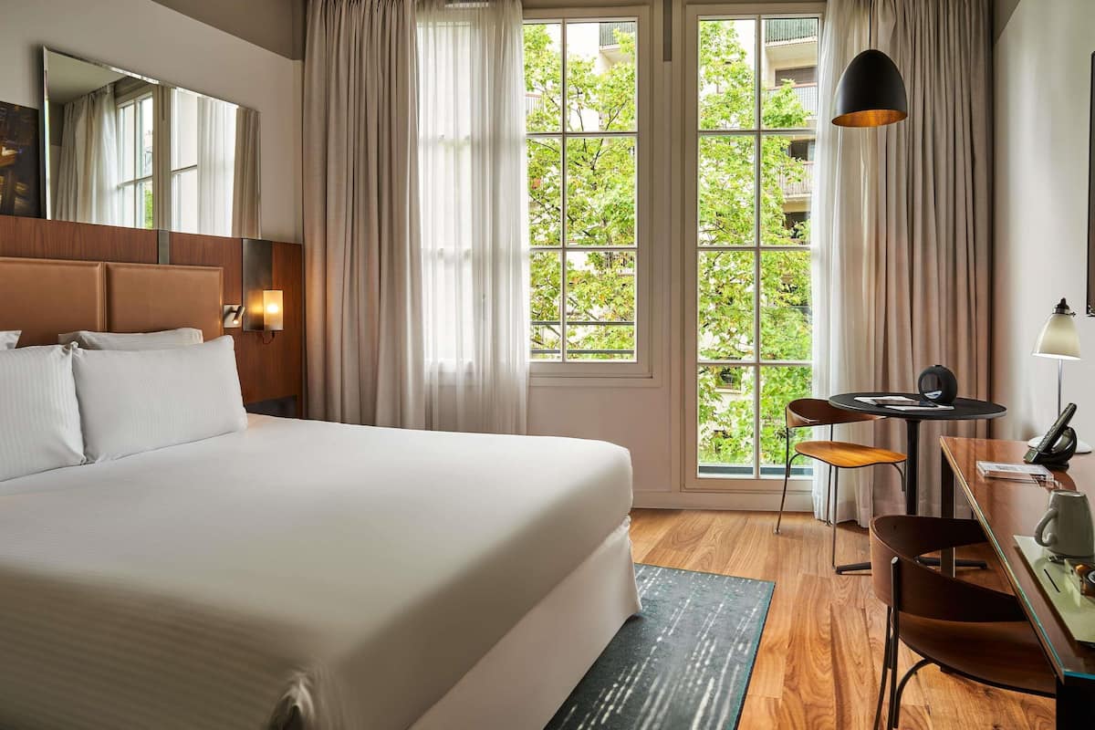 A hotel room with a large bed, a desk with a chair, a lamp, and a view of greenery through the window.