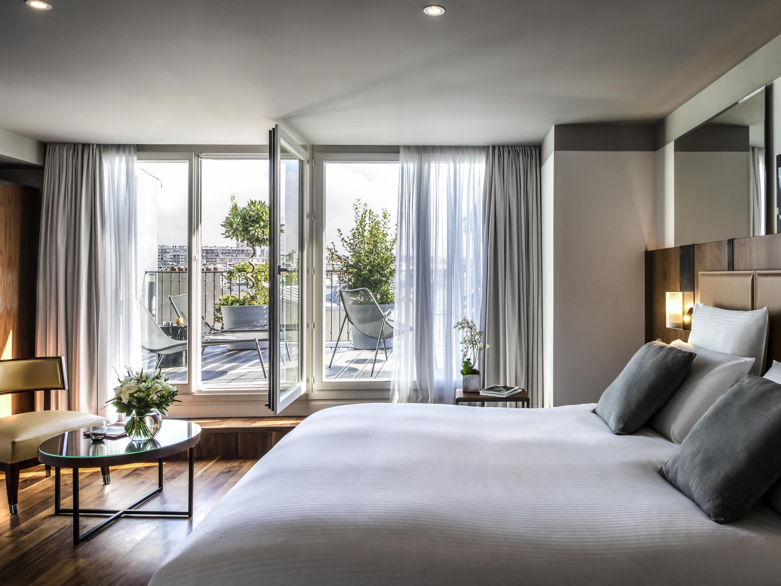 A modern hotel room with a large bed, a small table with a vase, a chair, and a view of the city through large windows.