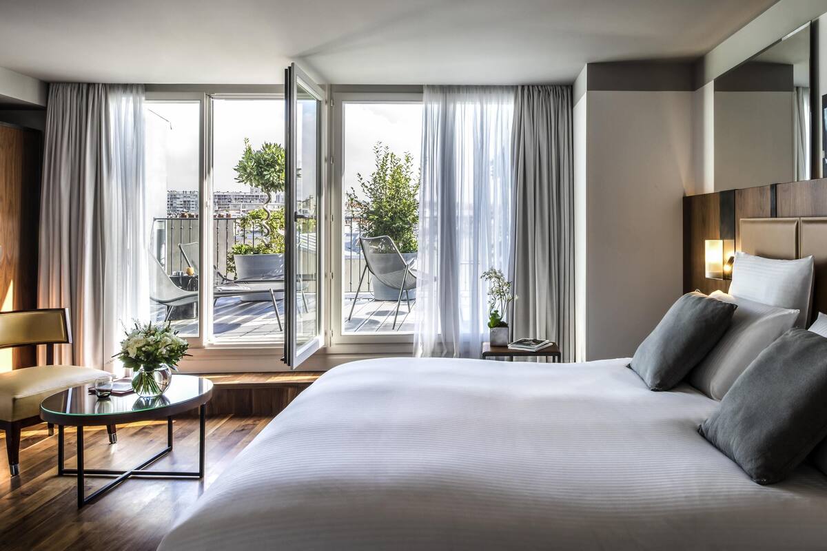 Une chambre d’hôtel moderne avec un grand lit, une petite table sur laquelle est posé un vase, une chaise et une vue sur la ville grâce à de grandes fenêtres.