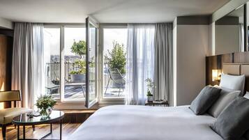 Una habitación de hotel moderna con una cama grande, una mesita con un florero, una silla y vistas a la ciudad a través de amplios ventanales.