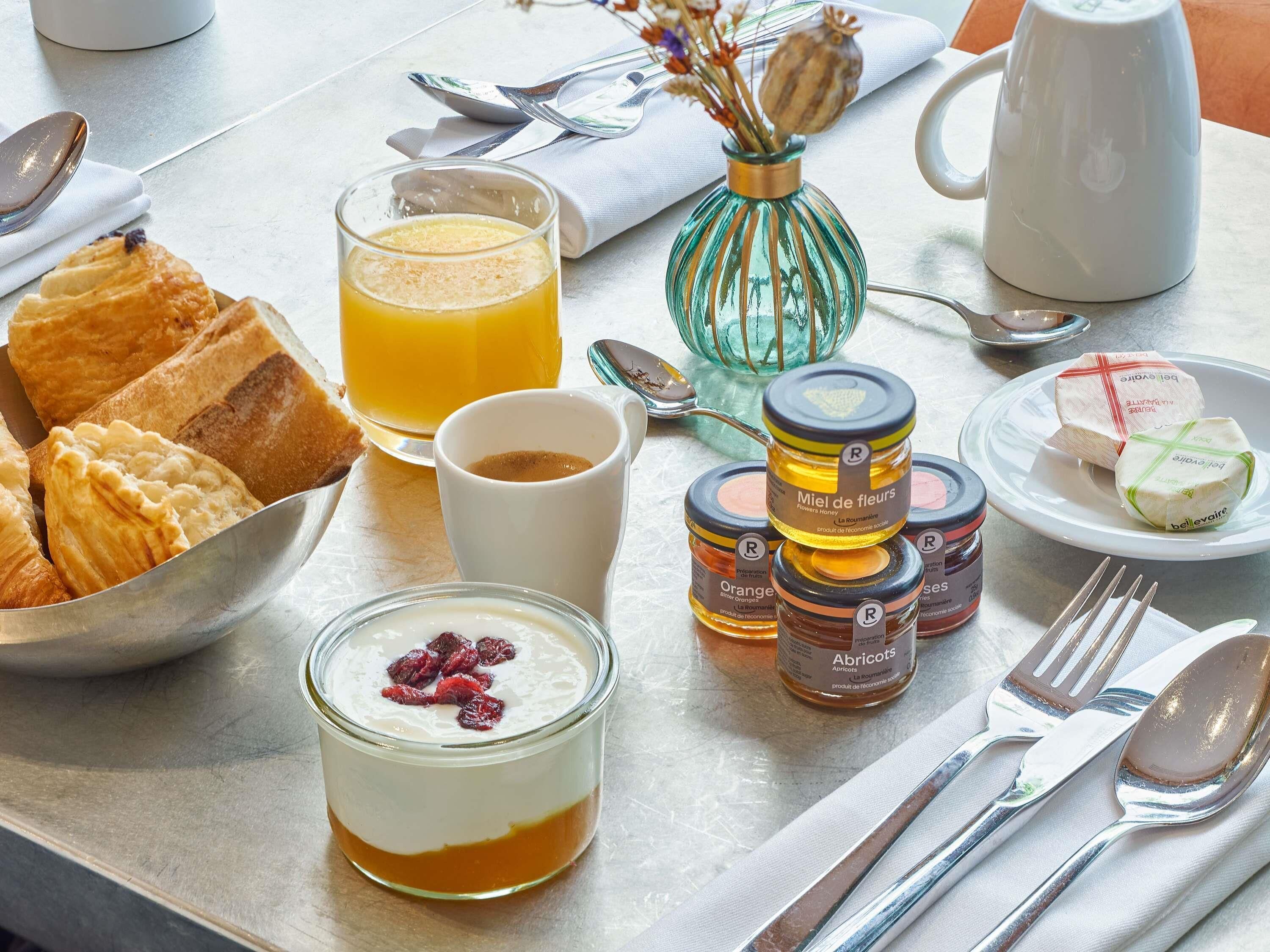 Una mesa de desayuno con pastelería, un vaso de zumo de naranja, tarros de miel, un parfait de yogur y una taza.