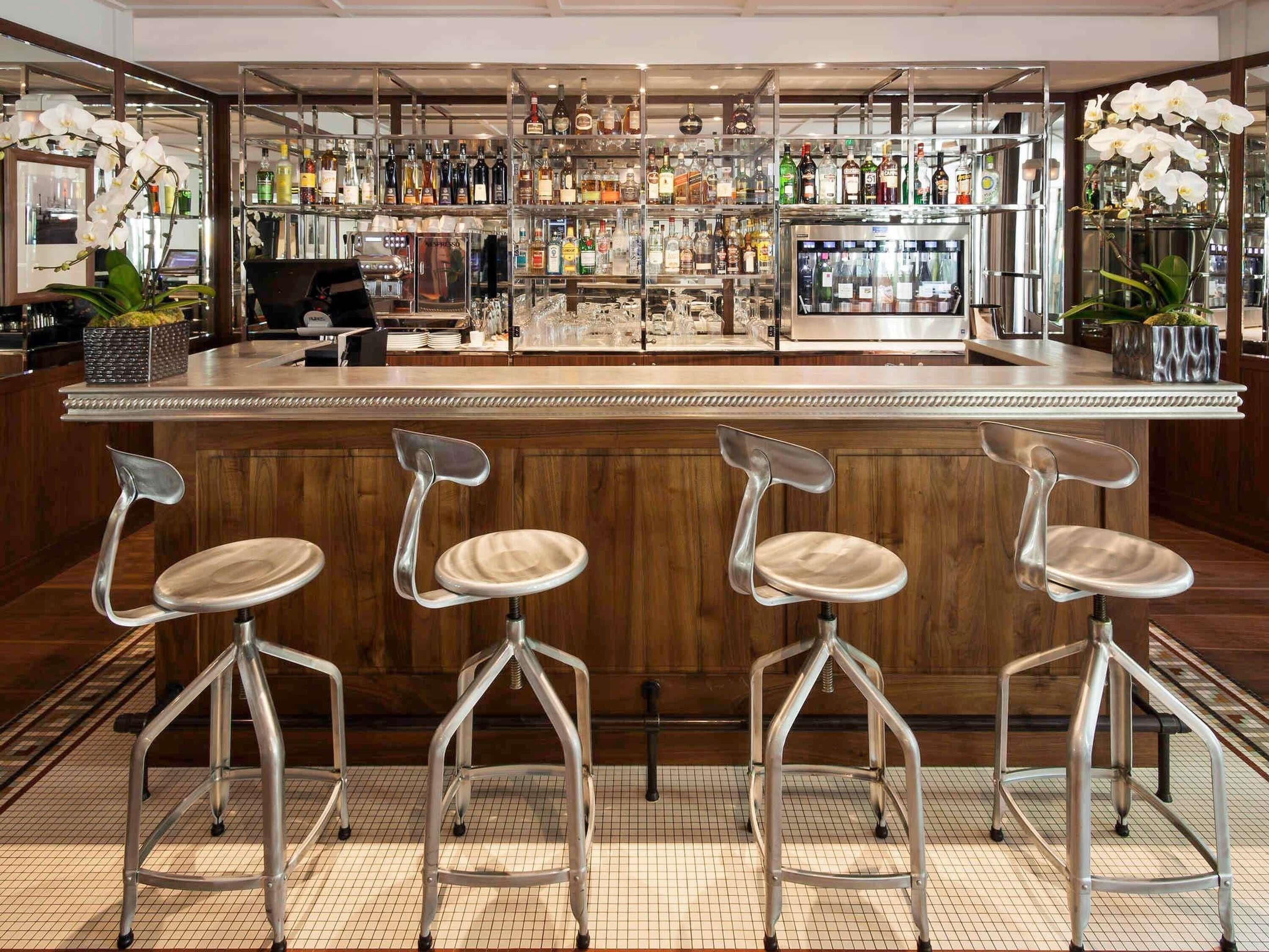 Zona de bar con mostrador de madera, taburetes metálicos y estantes llenos de botellas de licores.