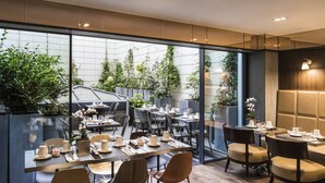 Moderni ravintola, jossa on lasiseinät, puiset pöydät ja tuolit. Ravintolasta avautuu näkymä vehreään puutarhaan ja lasiseen kasvihuoneeseen.