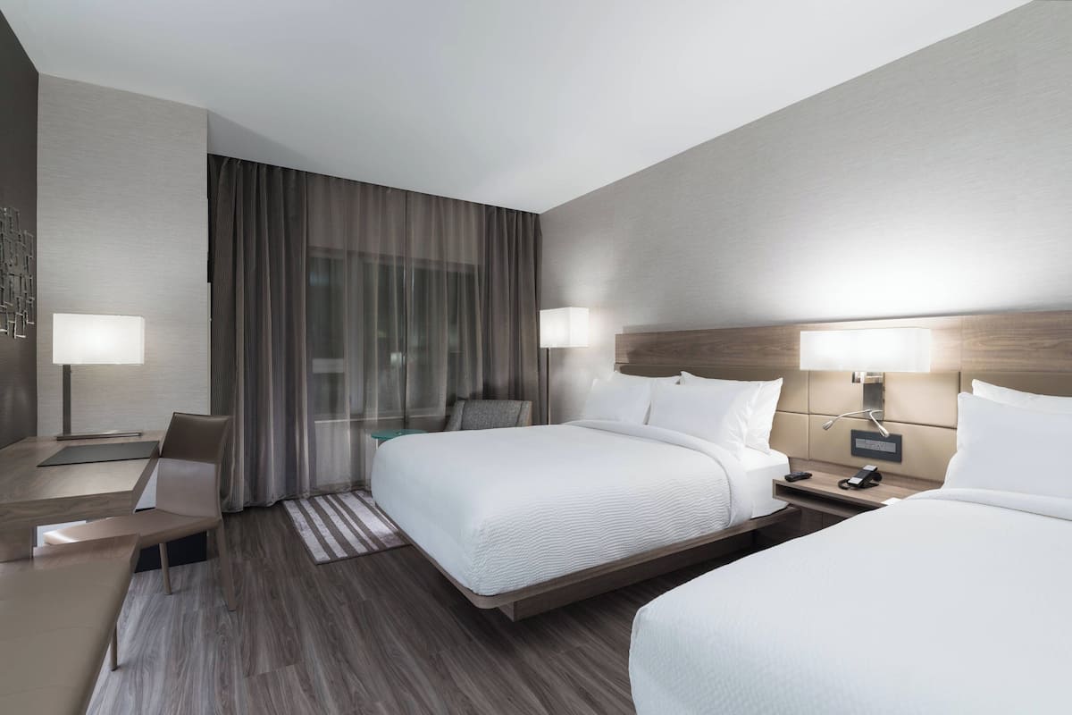 A modern hotel room with a large bed, a desk, and a chair. There is a window with curtains, a bedside table with a lamp, and a wall-mounted TV.