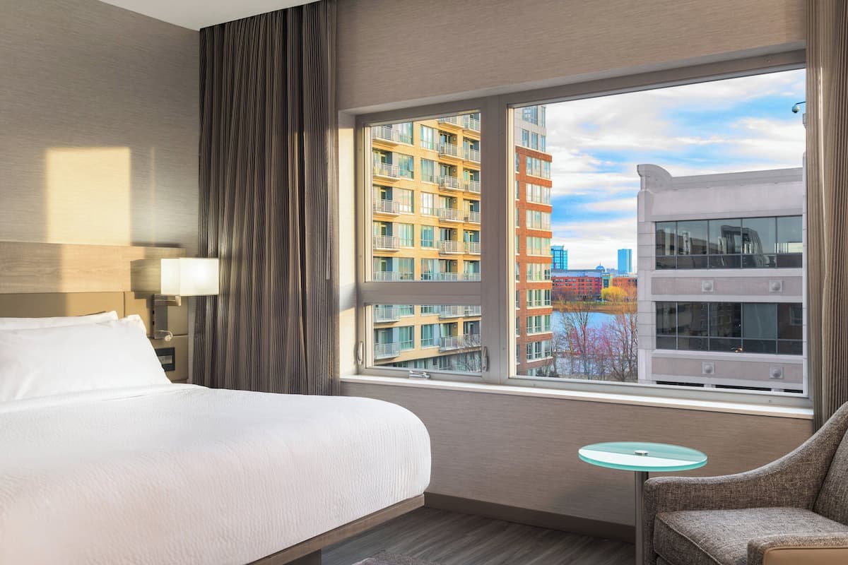 A hotel room with a large bed, a small round table, a chair, and a window offering a view of buildings and a body of water.