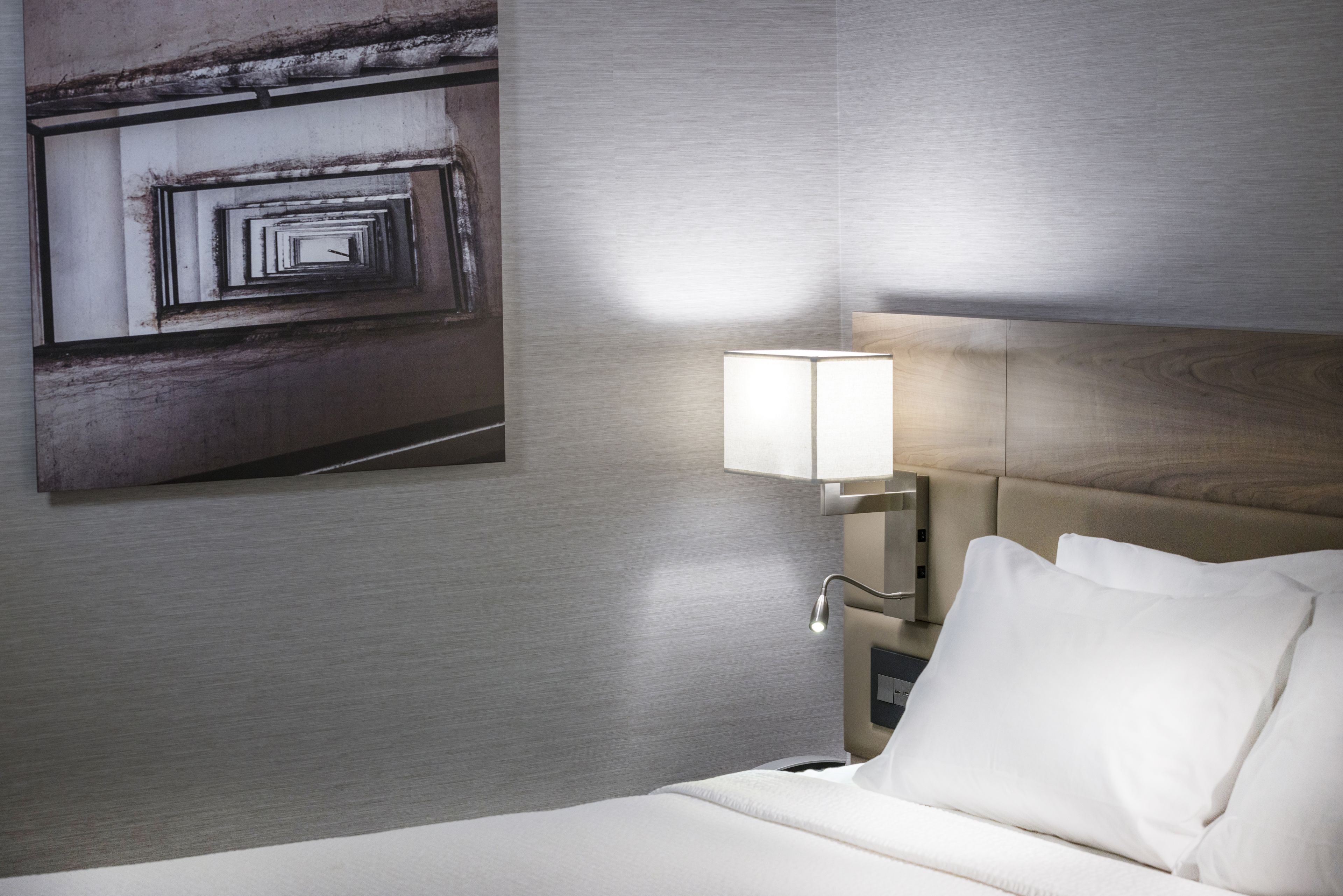 A hotel room with a modern design, featuring a neatly made bed, a wall-mounted lamp, and a large abstract painting.