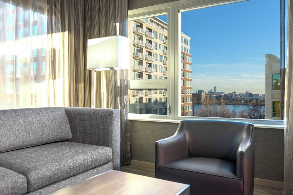 A hotel room with a grey sofa, a leather armchair, and a large window offering a city view.