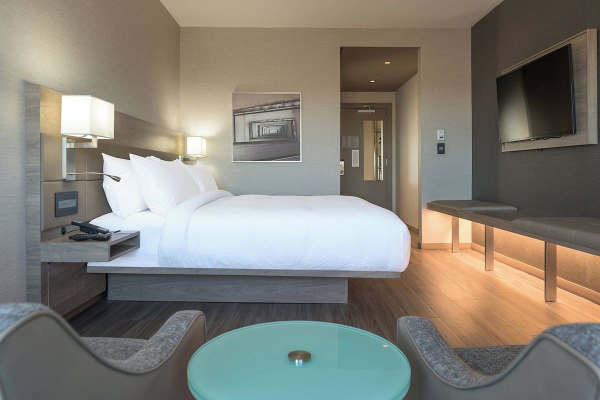 A modern hotel room with a large bed, bedside lamps, a flat-screen TV, a seating area with a table, and a wall-mounted artwork.