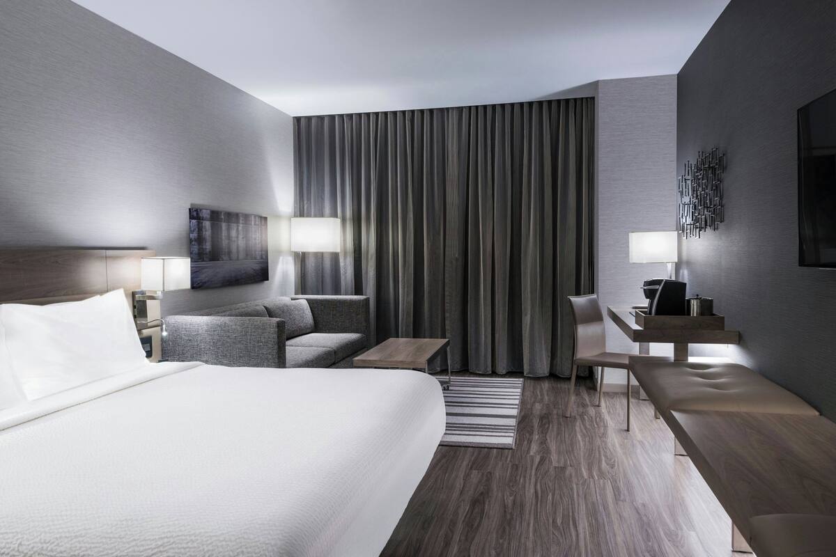 A modern hotel room with a large bed, a desk, a chair, a sofa, and a large window with curtains.