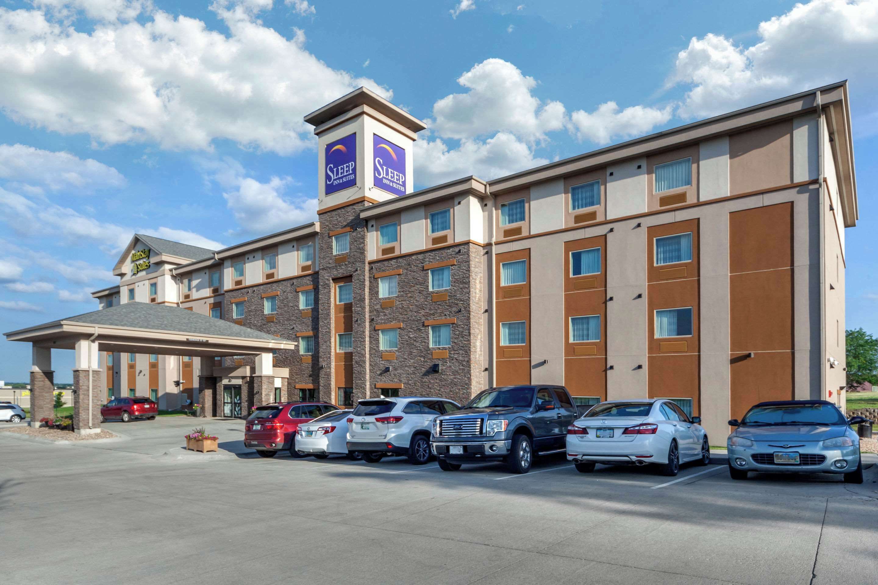 A hotel with a modern design, featuring a central entrance, multiple parking spaces, and a clear sky.