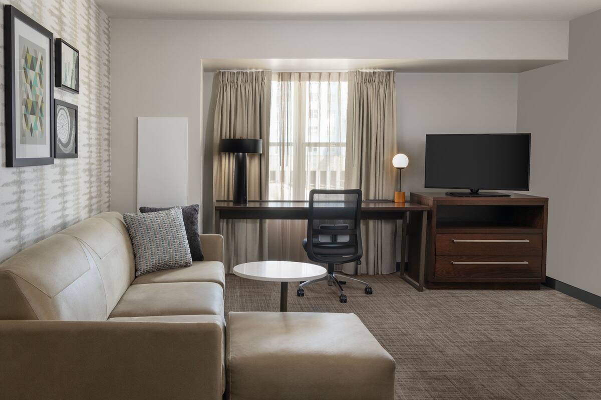 Ein Hotelzimmer mit einer Couch, einem runden Tisch, einem Schreibtisch mit Stuhl, einem Fernseher und einem Schrank.