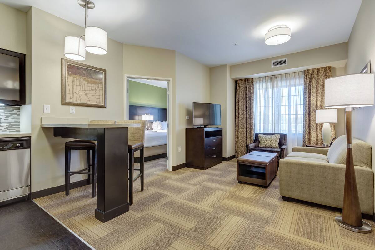 Habitación de hotel con kitchenette, un sofá, una mesa de centro, televisión y un amplio ventanal con cortinas.