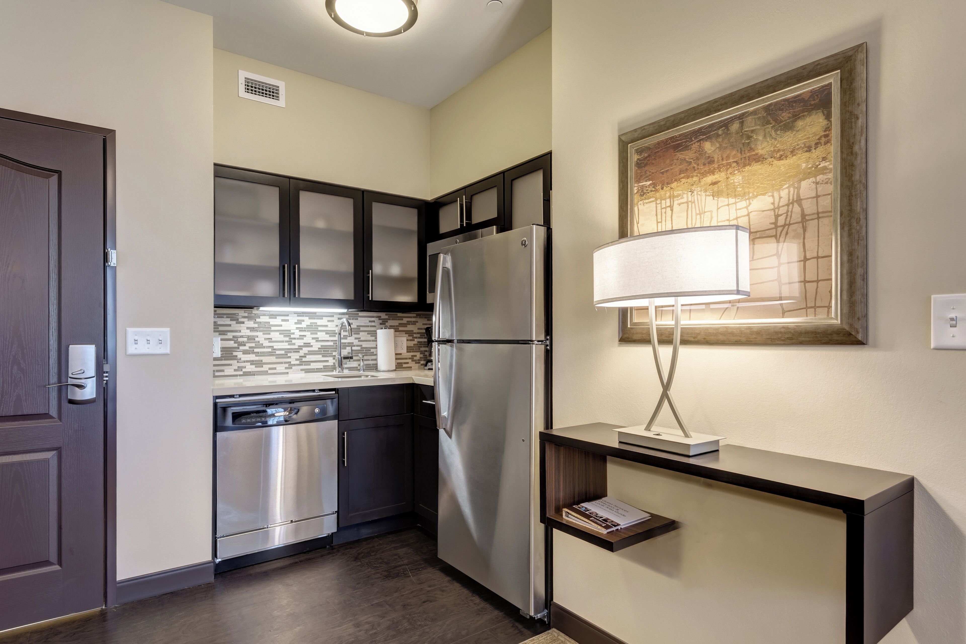 A modern kitchen with dark cabinets, stainless steel appliances, and a wall-mounted lamp.