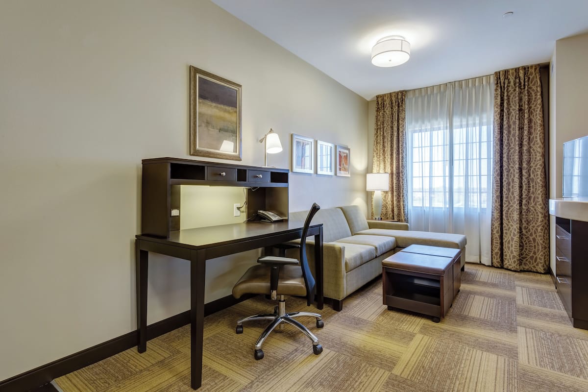 Una habitación de hotel con escritorio, silla de oficina, sofá y una mesita. En la pared hay cuadros enmarcados, una lámpara y una ventana con cortinas.