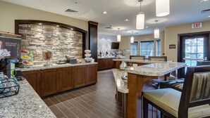 Una cocina moderna con encimeras de granito, armarios de madera y una pared decorativa de piedra. Hay taburetes altos y una zona de buffet con alimentos.
