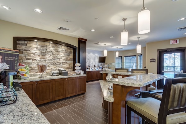 A modern kitchen with granite countertops, wooden cabinets, and a stone accent wall. There are bar stools and a buffet area with food items.