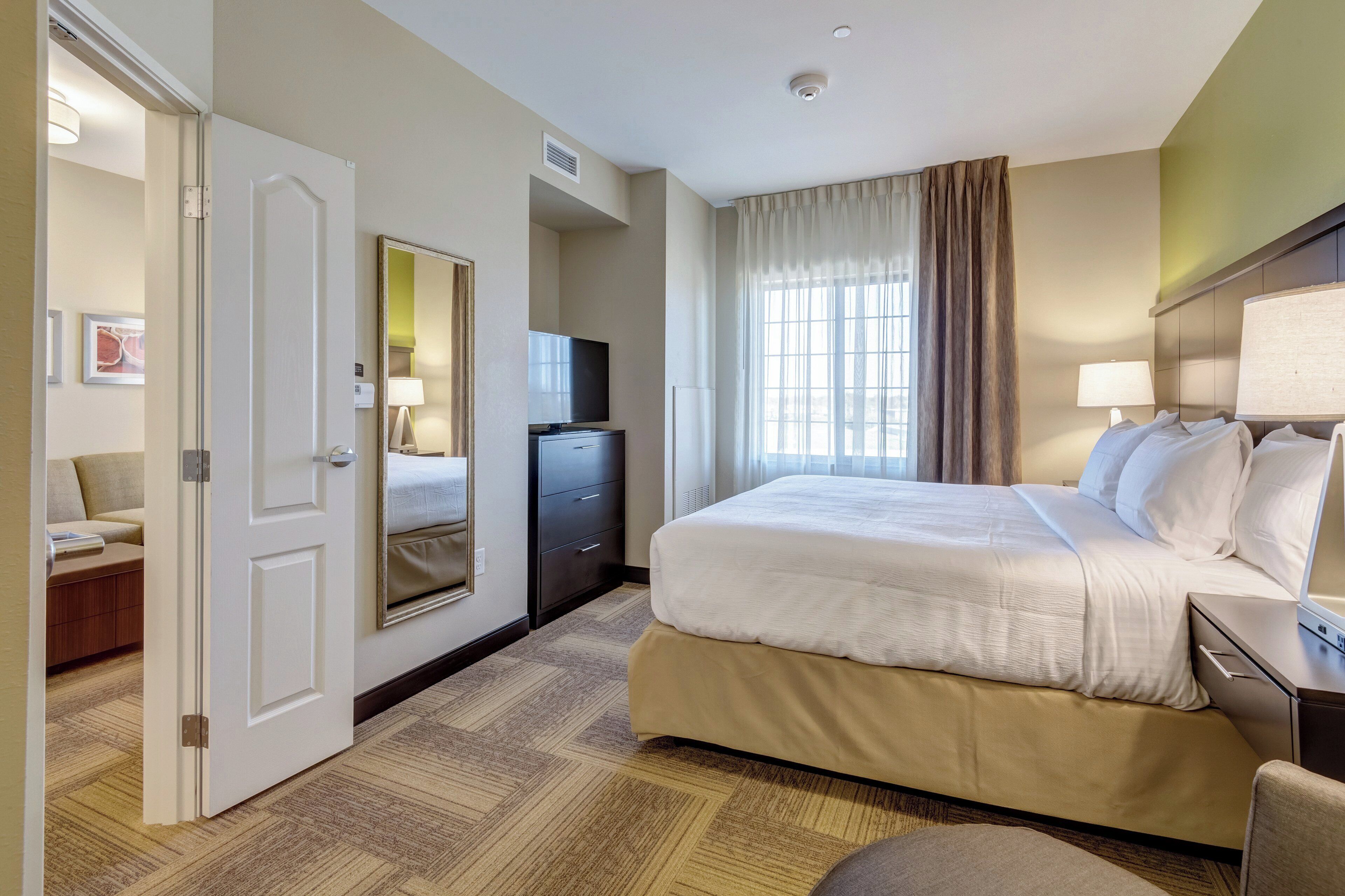 Ein Hotelzimmer mit Bett, Schreibtisch, Stuhl, Kommode, Fernseher und einem Fenster mit Vorhängen.