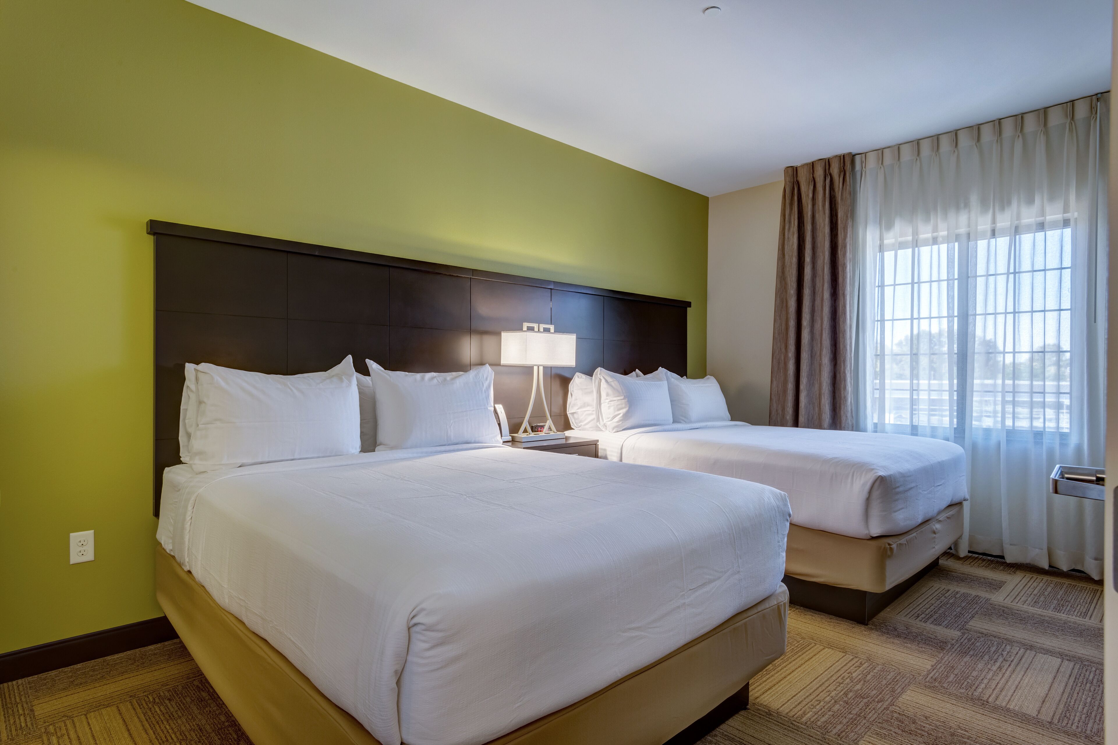 A hotel room with two beds, a nightstand with a lamp, a window with curtains, and a carpeted floor.