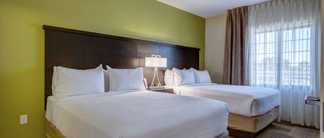 A hotel room with two beds, a nightstand with a lamp, a window with curtains, and a carpeted floor.