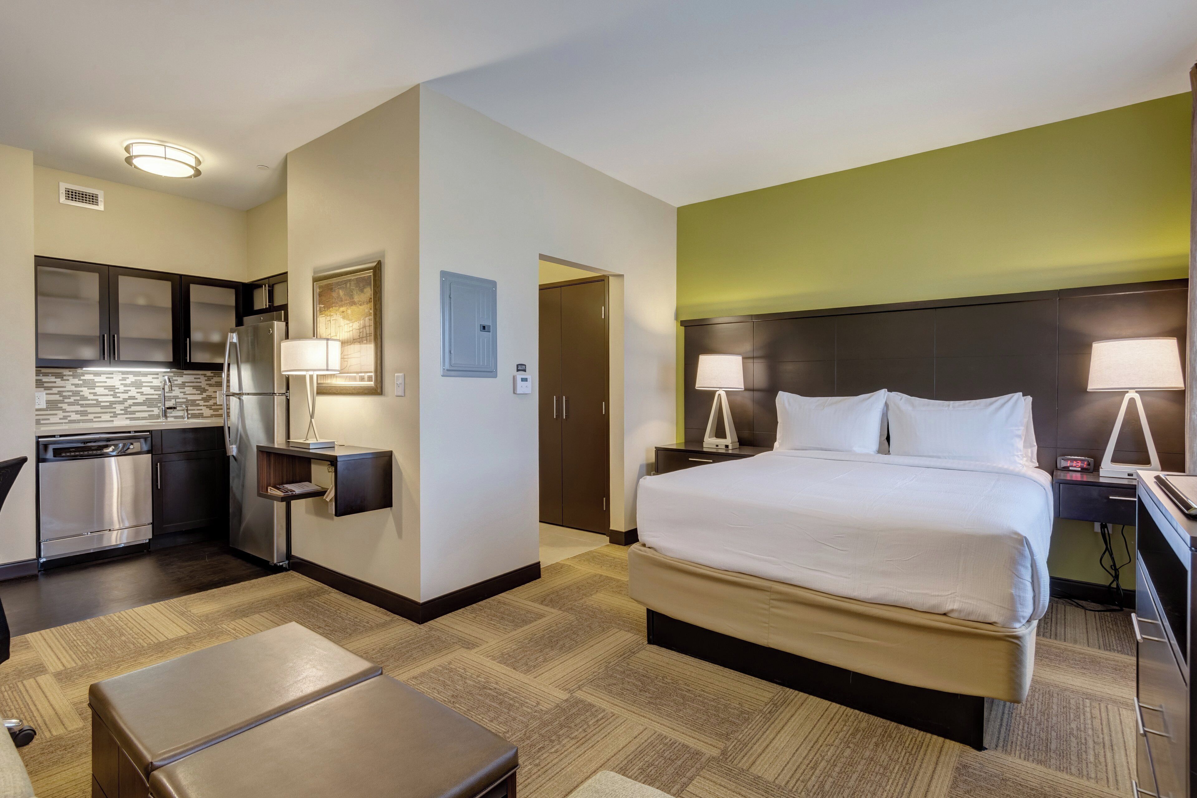 A hotel room with a bed, bedside lamps, a desk, a chair, a small table, a refrigerator, a microwave, and a kitchen area with cabinets and a dishwasher.