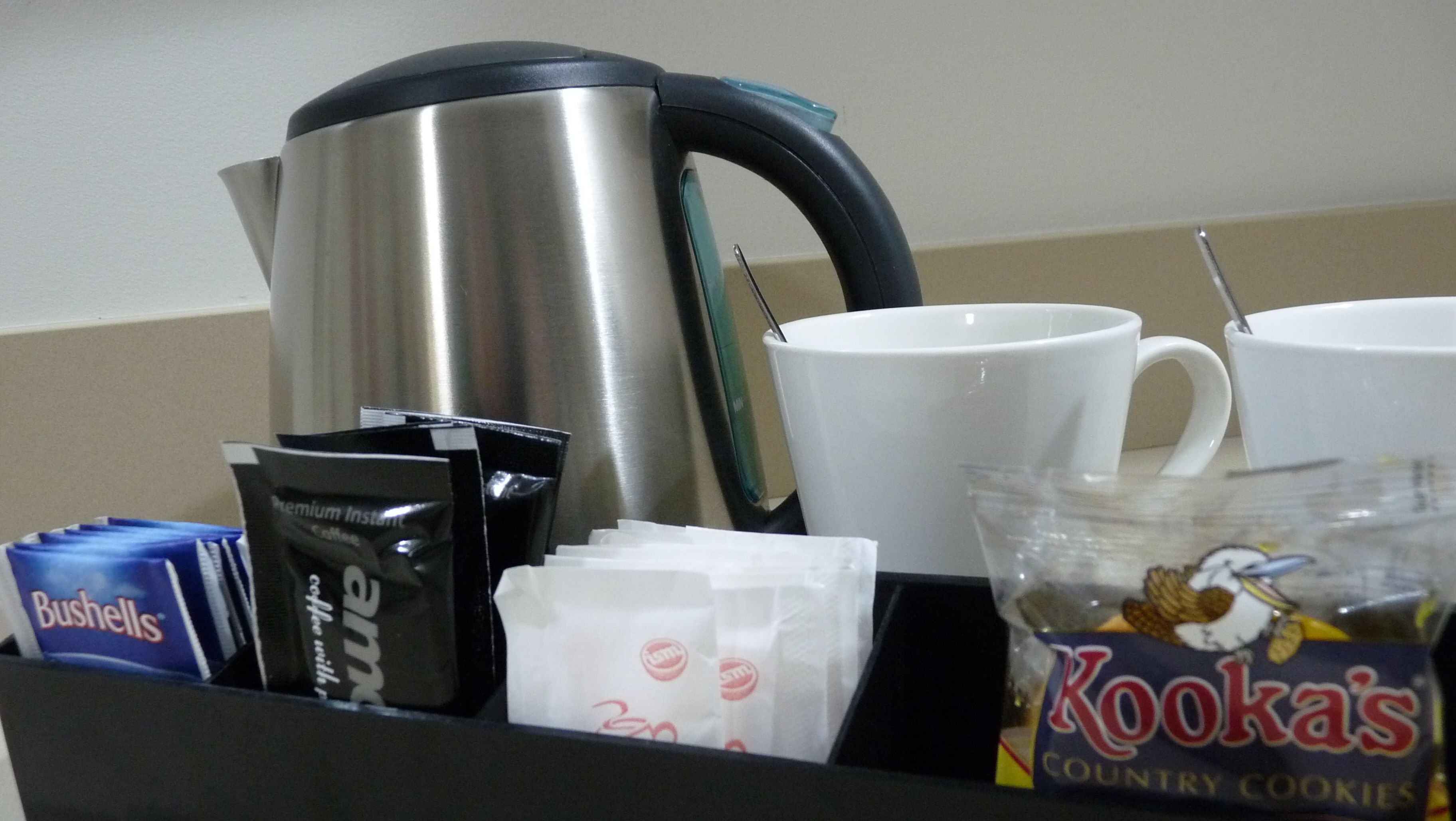 Una bandeja con una tetera de acero inoxidable, tazas blancas, un paquete de galletas Kook's country, un paquete de café Bushells y un sobre de café instantáneo.