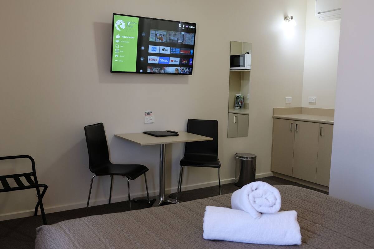 Habitación de hotel con cama, escritorio, dos sillas, televisor montado en la pared, microondas, cesto de basura y un mueble pequeño.