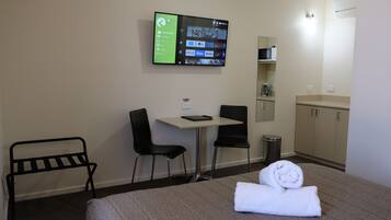 Quarto de hotel com uma cama, uma escrivaninha, duas cadeiras, uma TV fixada na parede, um micro-ondas, uma lixeira e um pequeno armário.