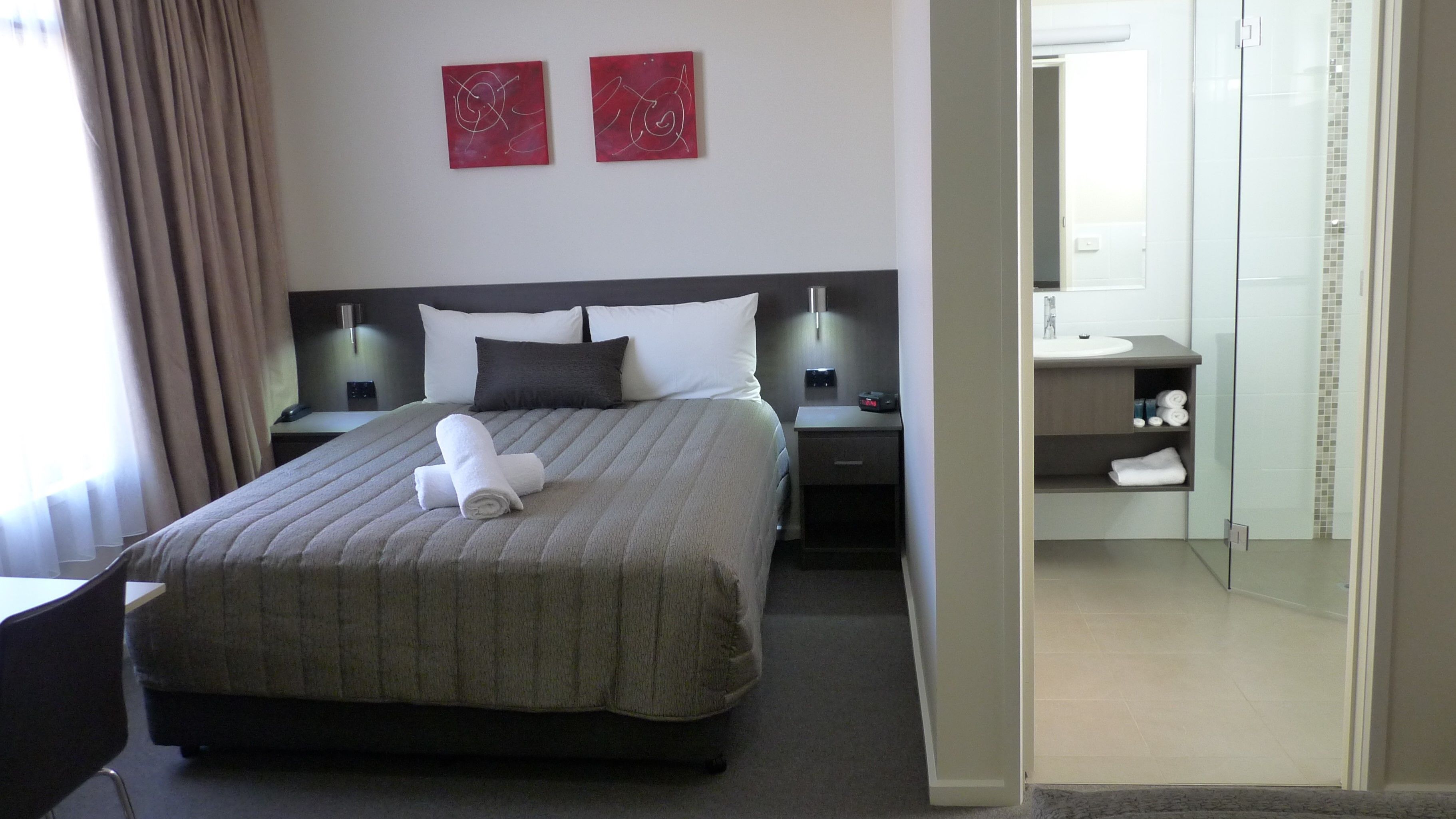 A hotel room with a bed, two bedside tables, a nightstand, a chair, and a bathroom visible through an open door.