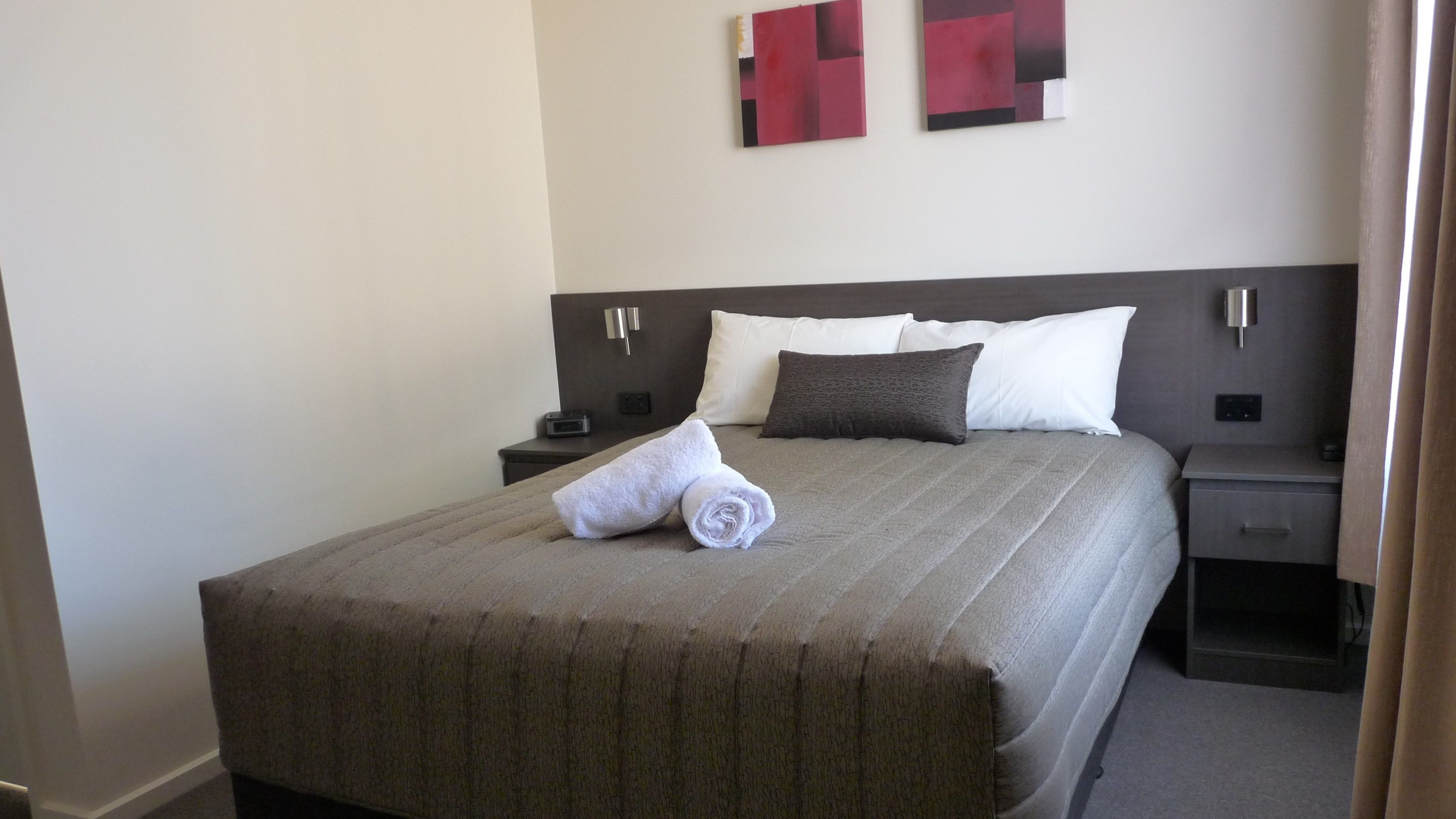 A neatly made bed with a brown bedspread, white and dark gray pillows, and a rolled white towel on the side. There are two bedside tables with lamps and a wall-mounted light.