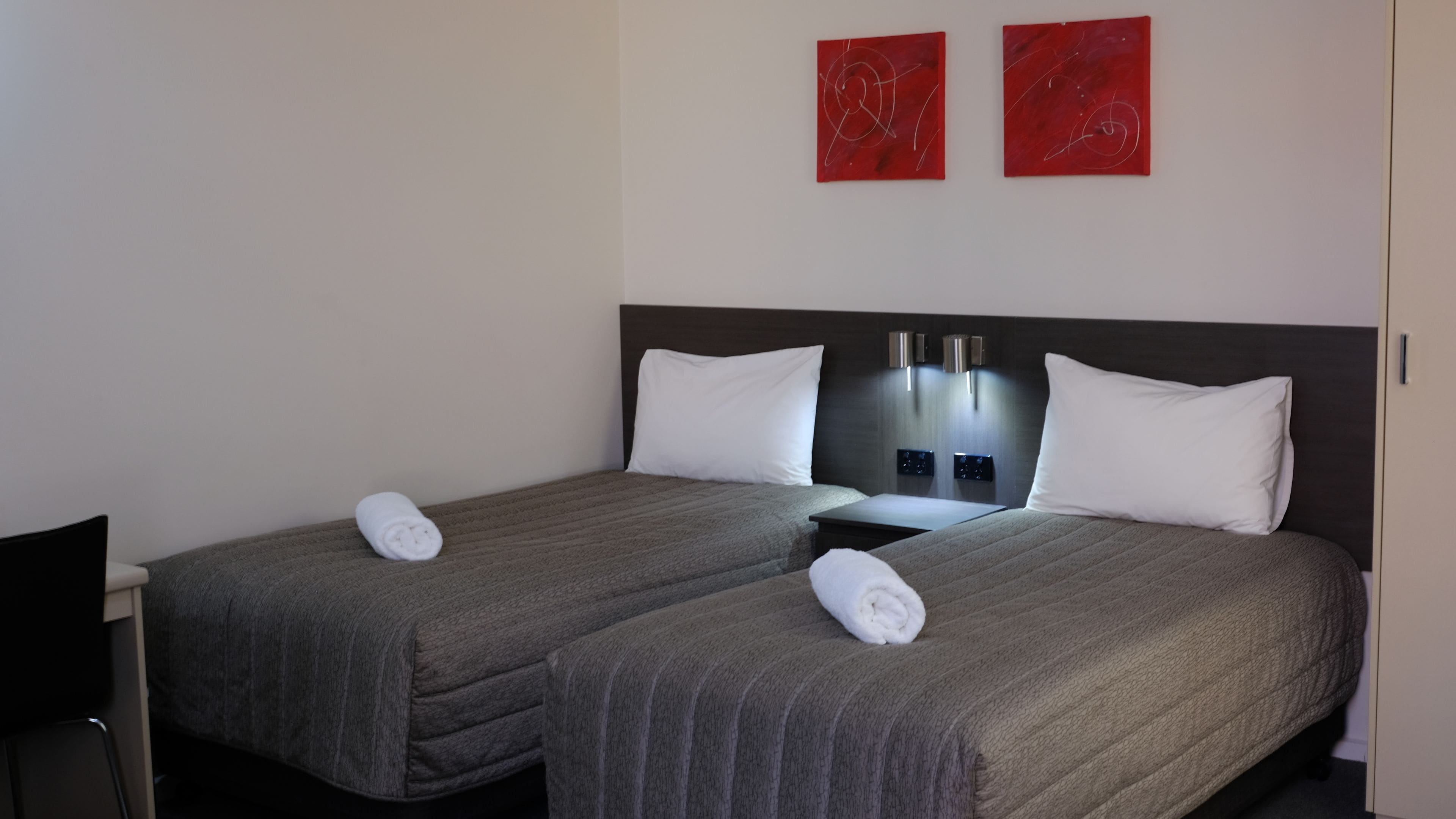 Habitación de hotel con dos camas, cada una con cubrecama gris y almohadas blancas. Dos cuadros abstractos rojos decoran la pared sobre las camas.