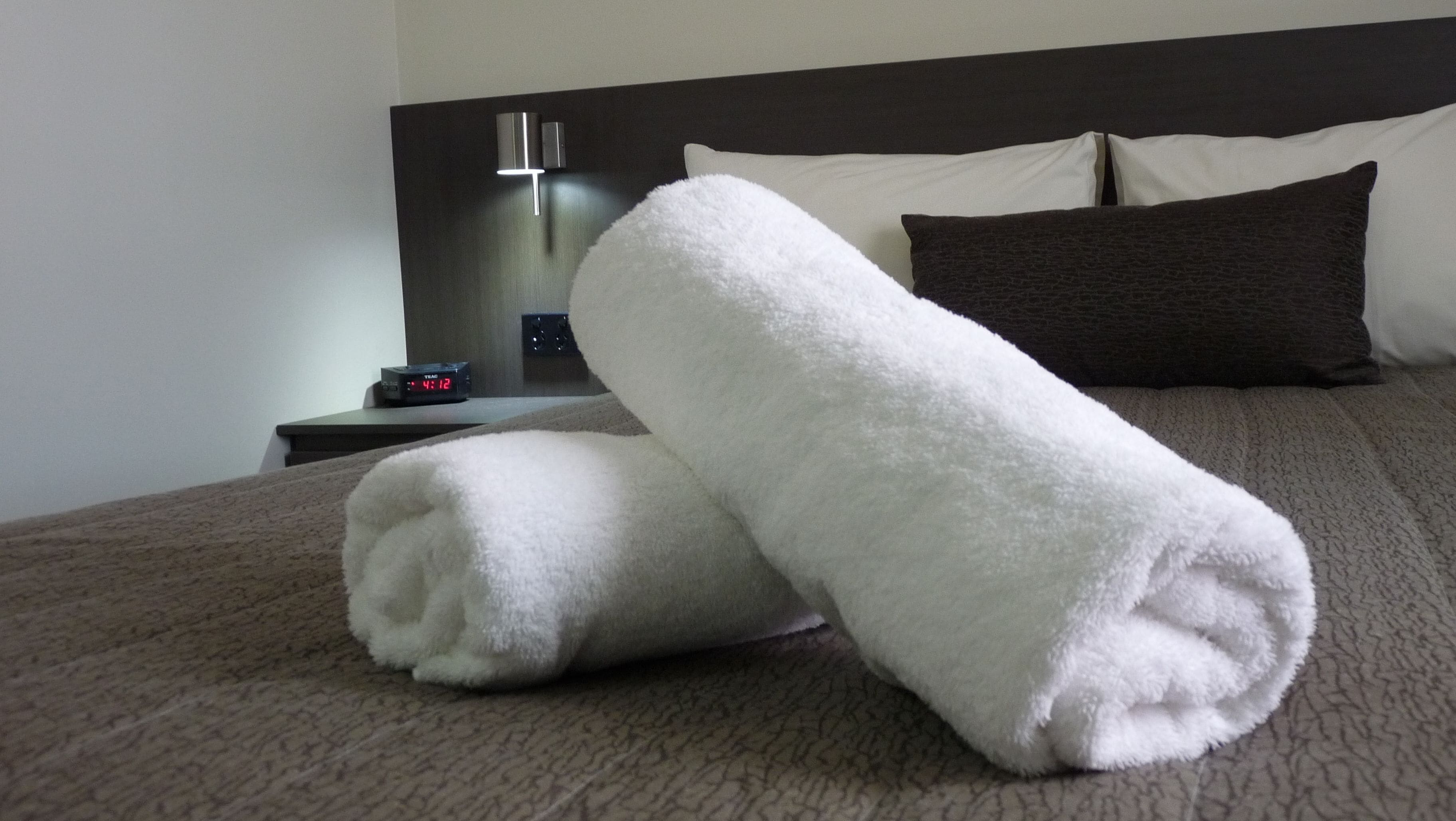 A neatly made bed with white towels, a dark headboard, and a bedside table with a clock.