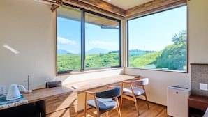 Una habitación con un ventanal grande, un escritorio de madera, dos sillas, una kitchenette pequeña y vistas a un paisaje verde.