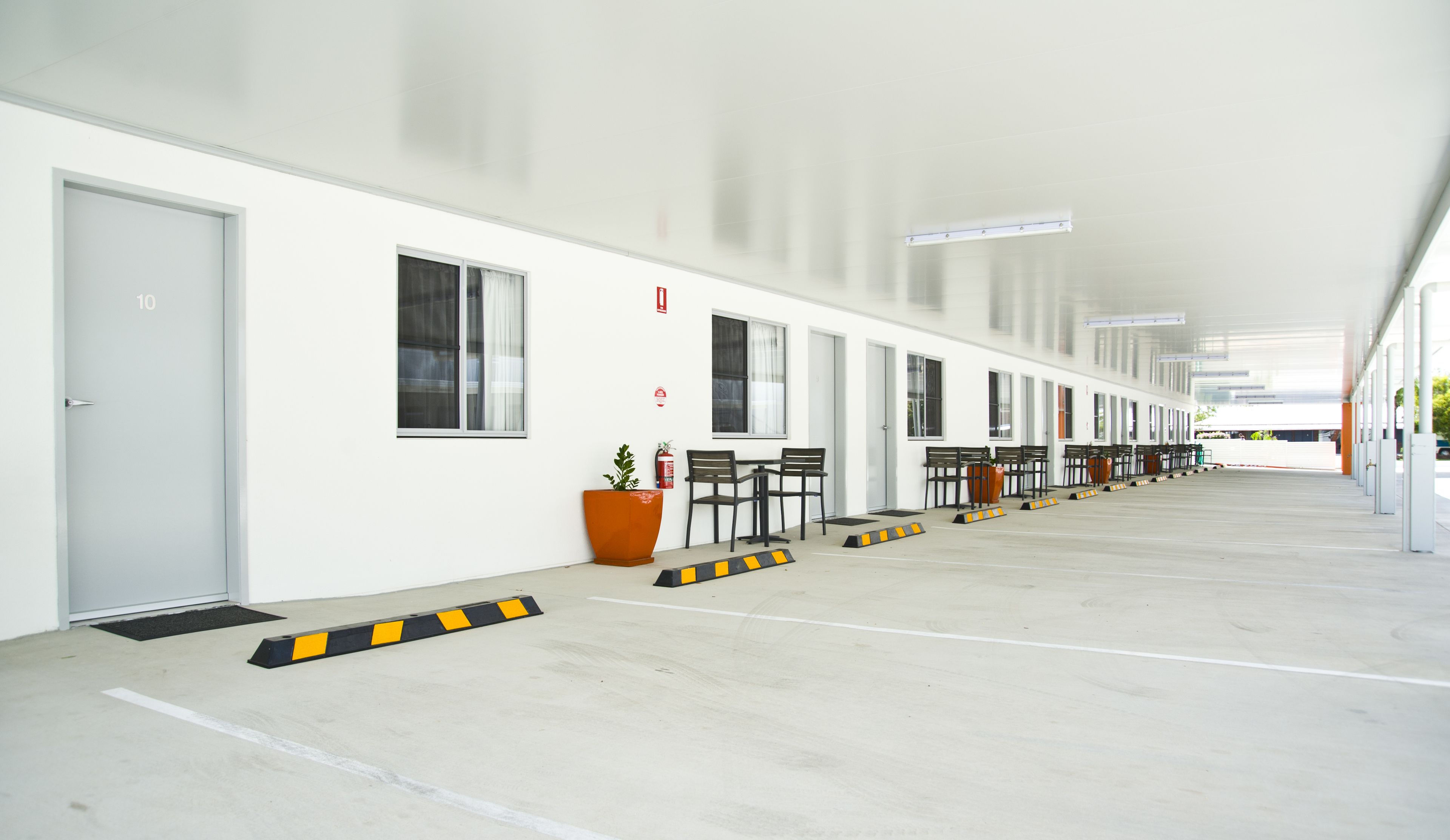 A clean, modern hotel corridor with white walls, a row of doors numbered 10, and a series of tables and chairs along the side.