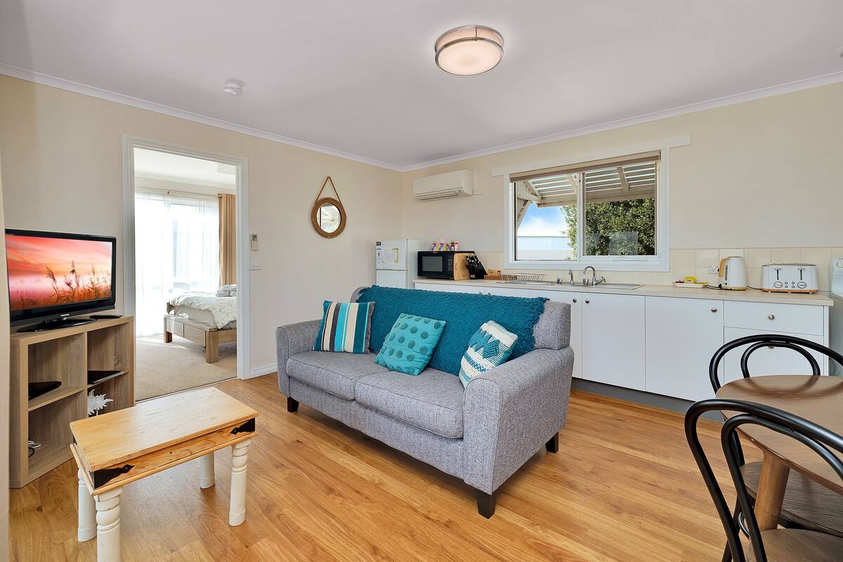 A living room with a grey sofa, wooden coffee table, and a television. There is a kitchen area with a microwave, toaster, and sink.