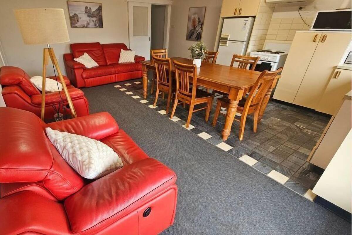 A compact living space with a red sofa, a dining table with chairs, a kitchen area with cabinets, and a small rug.