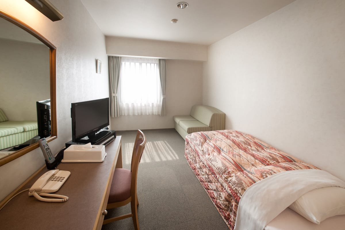 Una camera d'albergo con un letto, una scrivania con il telefono, una televisione e una finestra con tende.