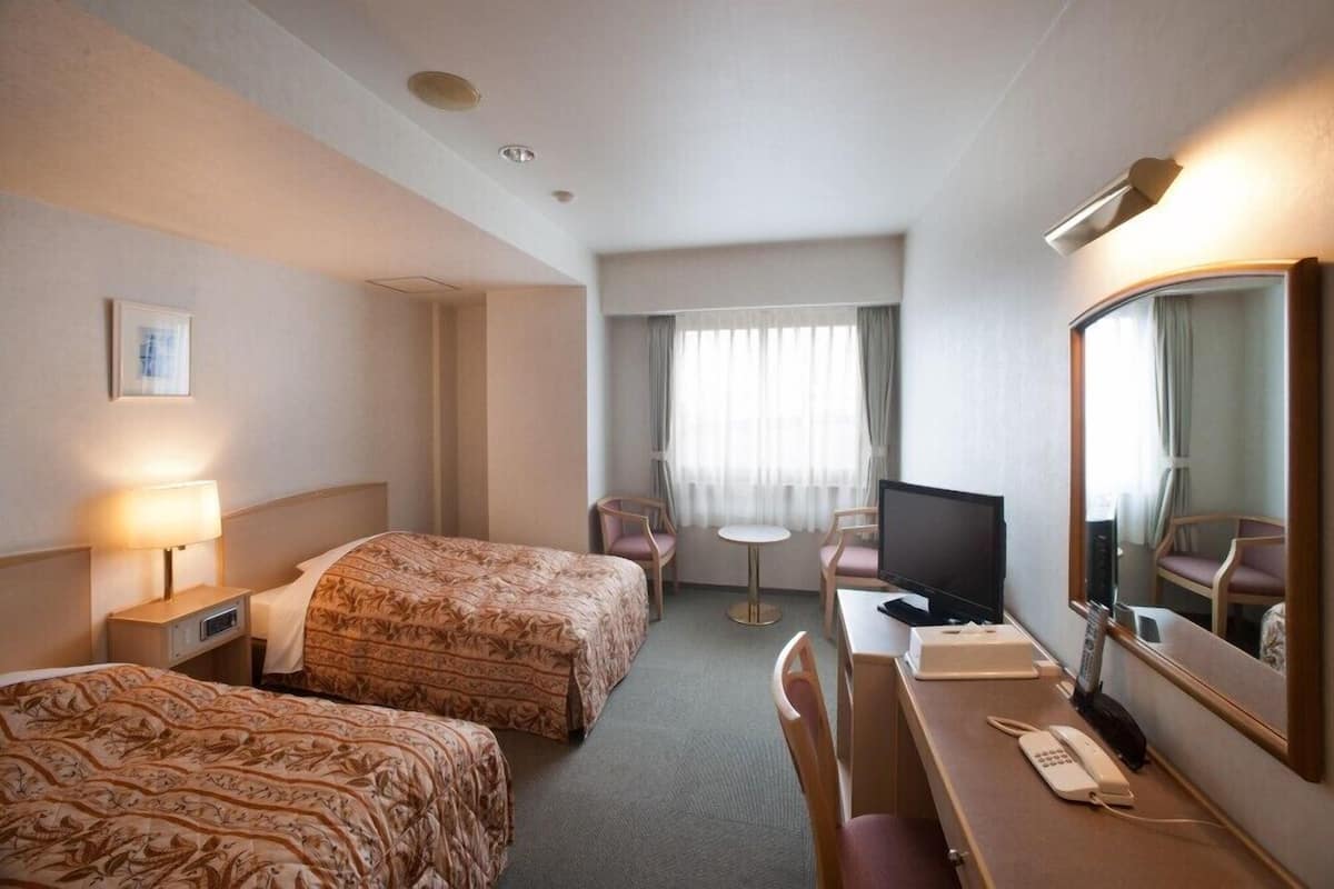 Una camera d'albergo con due letti, una scrivania con telefono e televisione, un tavolino e una finestra con tende.