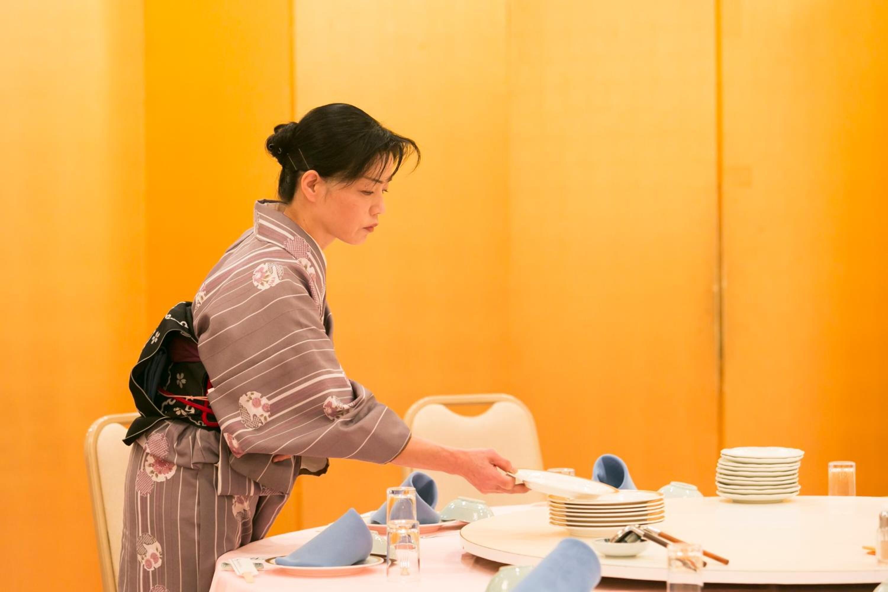 A person seated at a table with plates, bowls, and chopsticks, in a room with orange walls.