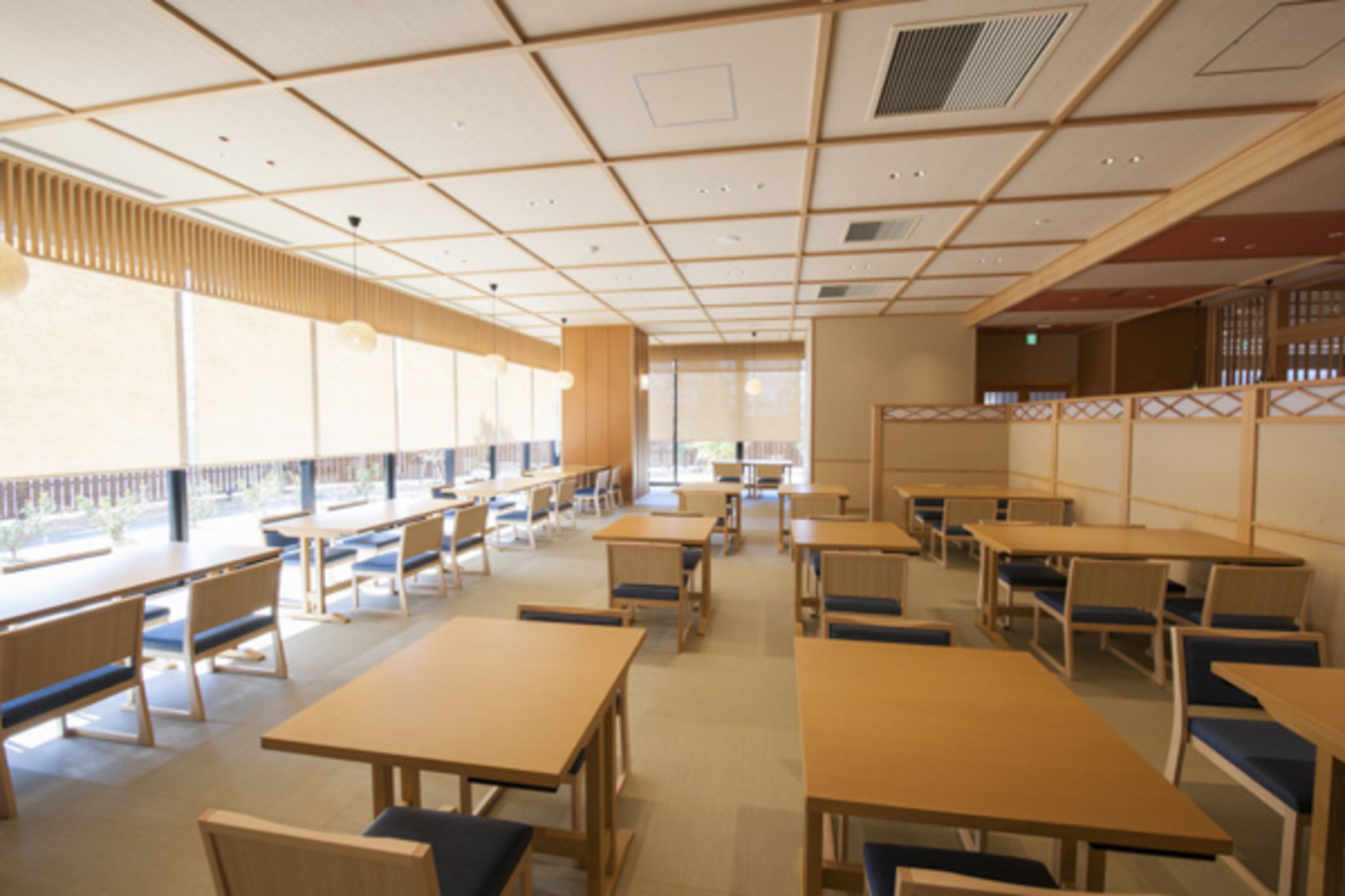 A spacious dining area with multiple tables and chairs, large windows providing natural light, and a traditional Japanese-style partition wall.