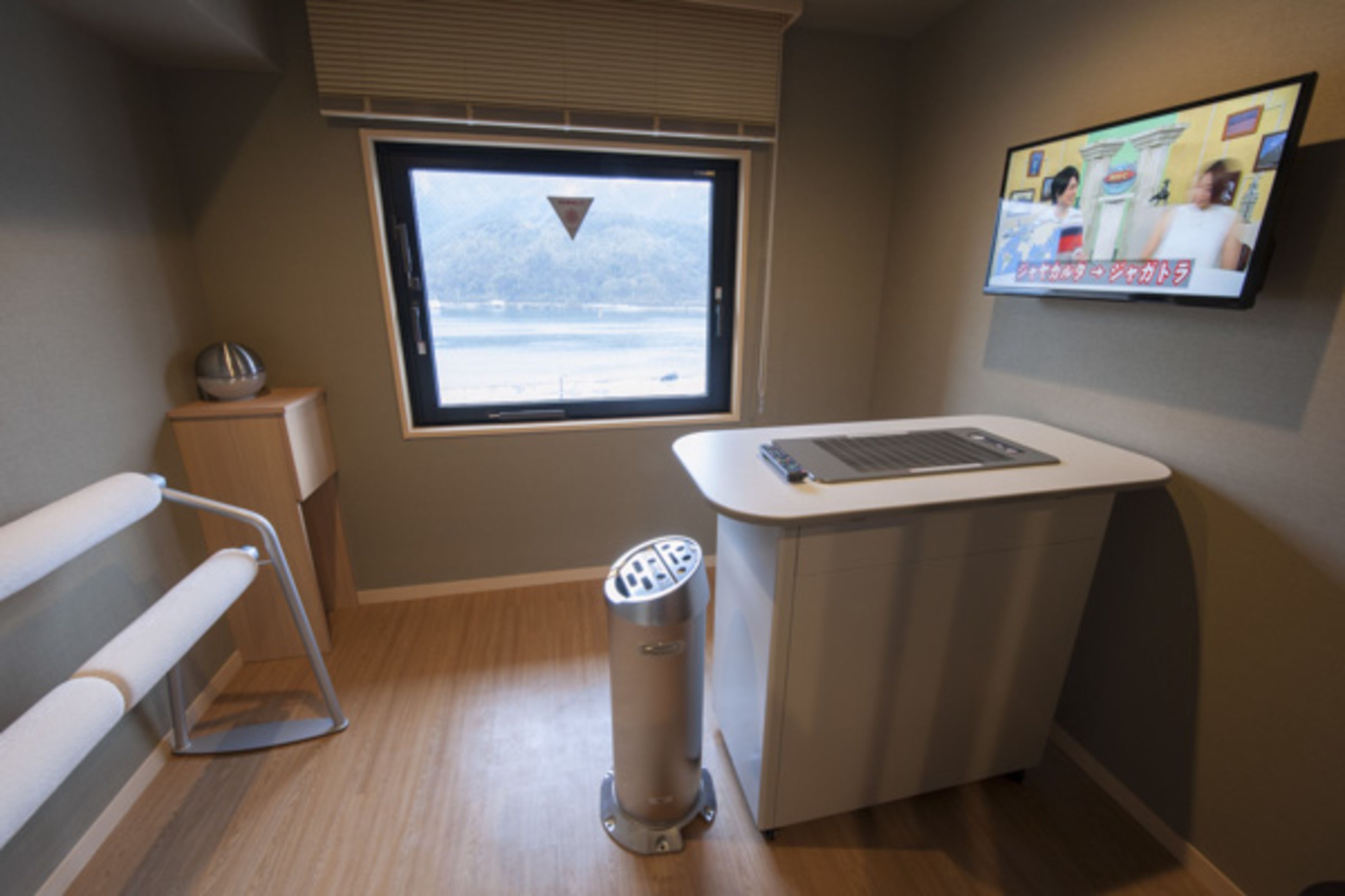Een kamer met een raam, een bureau, een stoel en een aan de muur bevestigde televisie.