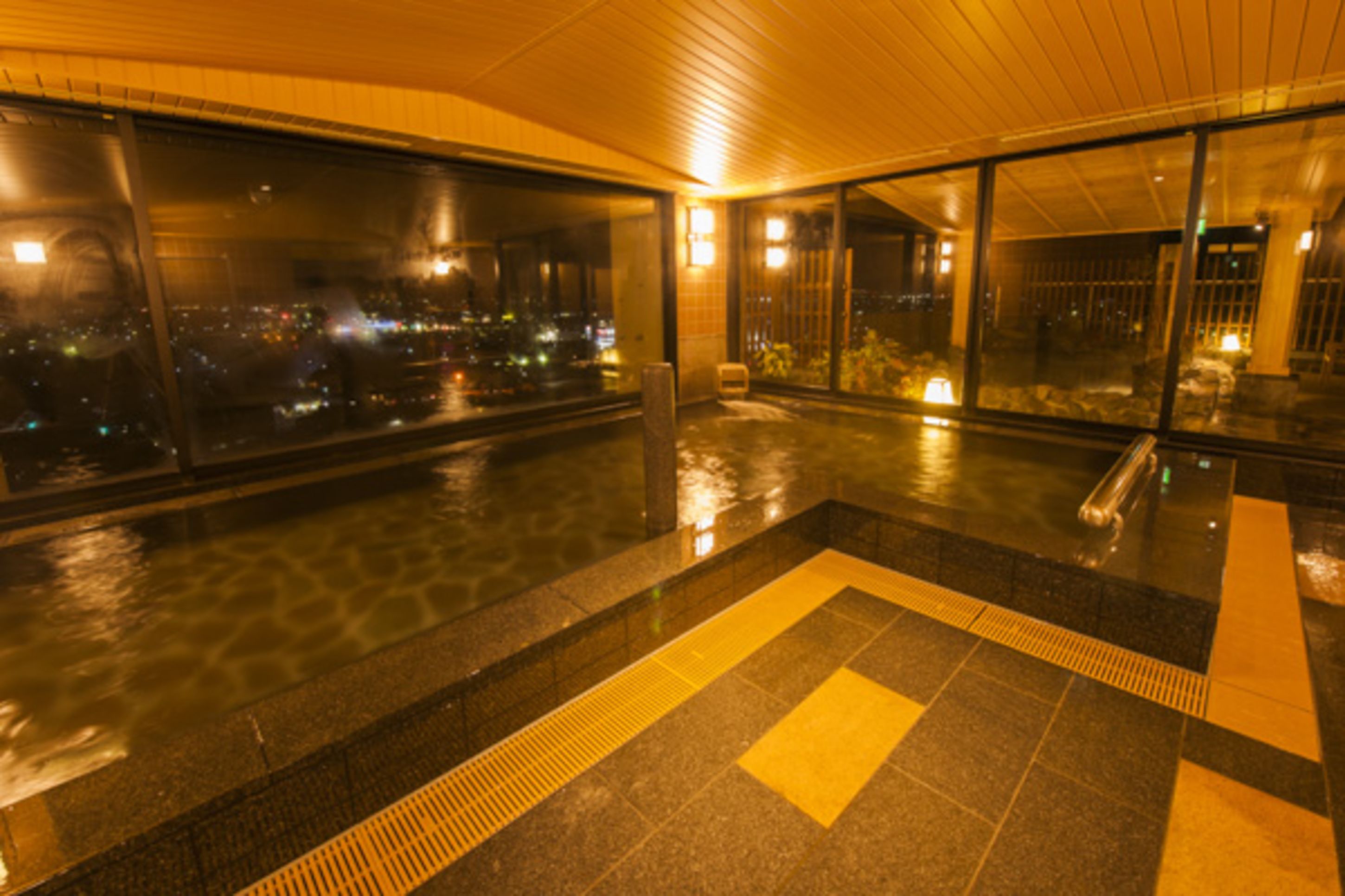 Un área de piscina interior moderna con amplios ventanales, piso de baldosas y una vista panorámica de la ciudad de noche.