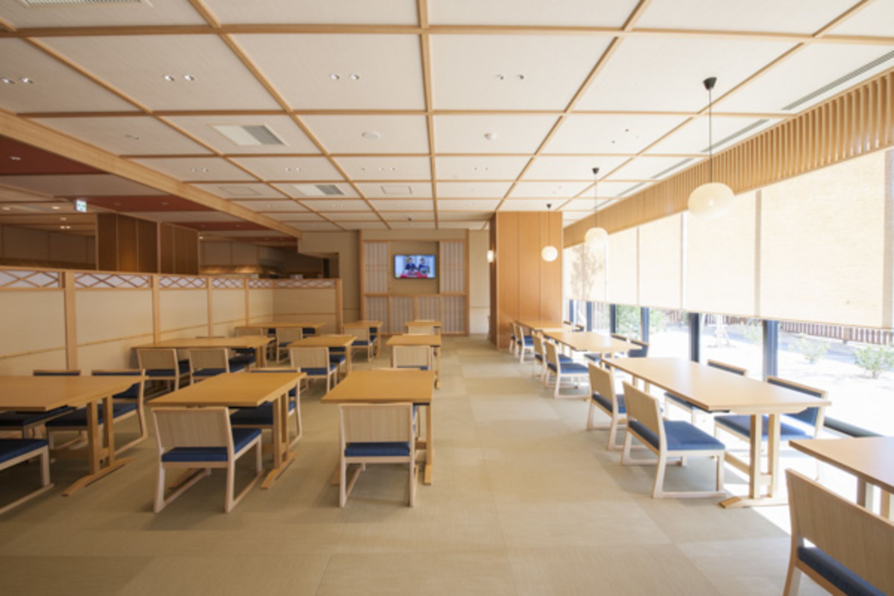 A spacious dining area with wooden tables and chairs, a television screen, and large windows providing natural light.