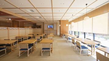 A spacious dining area with wooden tables and chairs, a television screen, and large windows providing natural light.