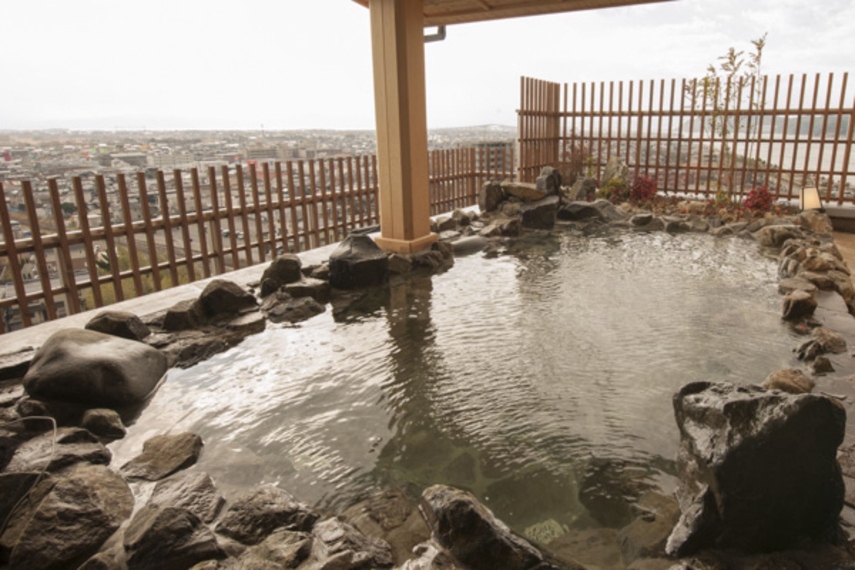 A stone water feature with a wooden pavilion and a metal fence overlooking a cityscape.