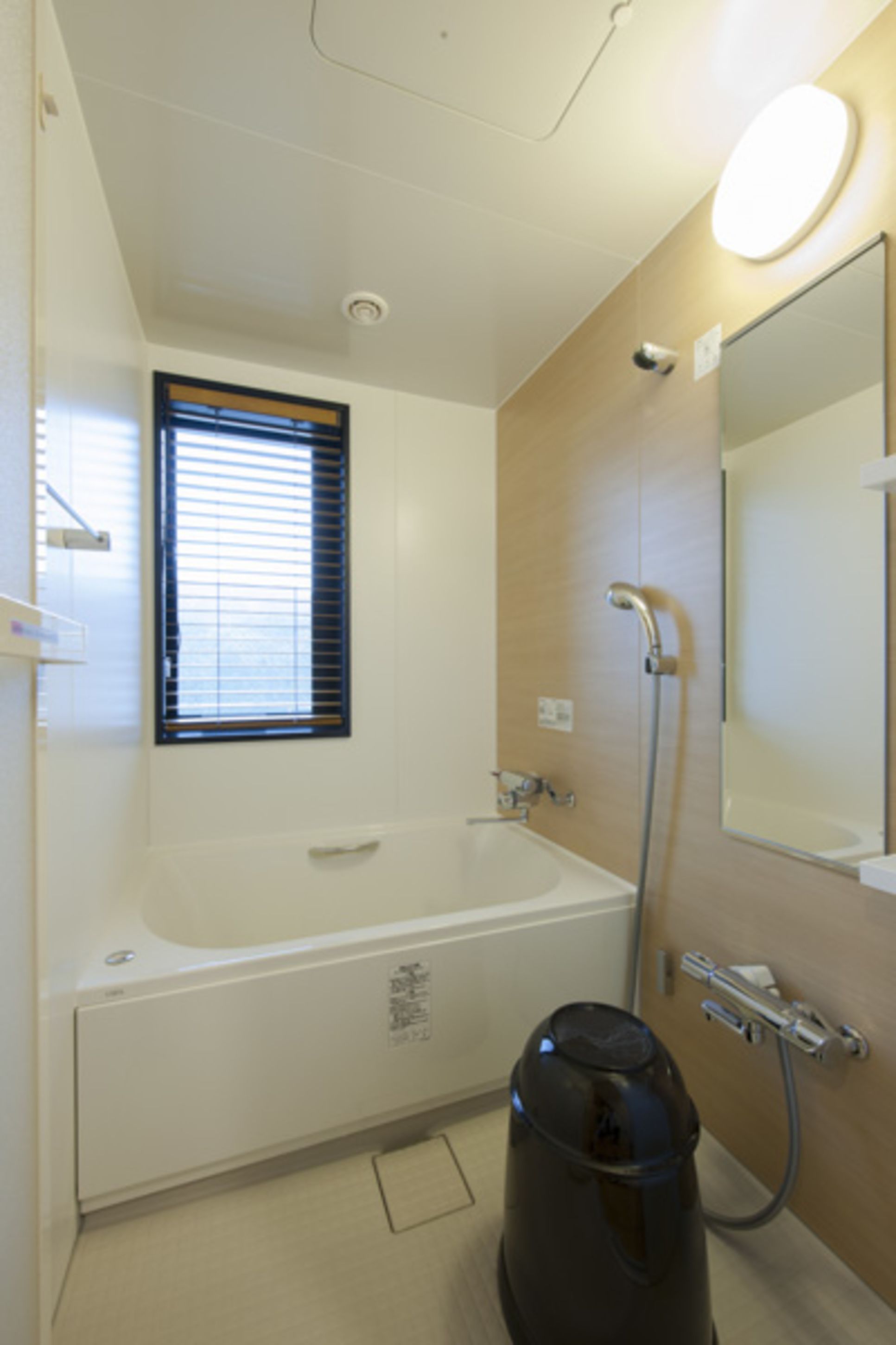 A bathroom with a bathtub, a window with blinds, a shower, a mirror, and a vacuum cleaner.