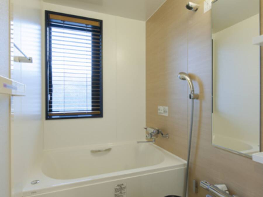 A bathroom with a bathtub, a window with blinds, a shower, a mirror, and a vacuum cleaner.